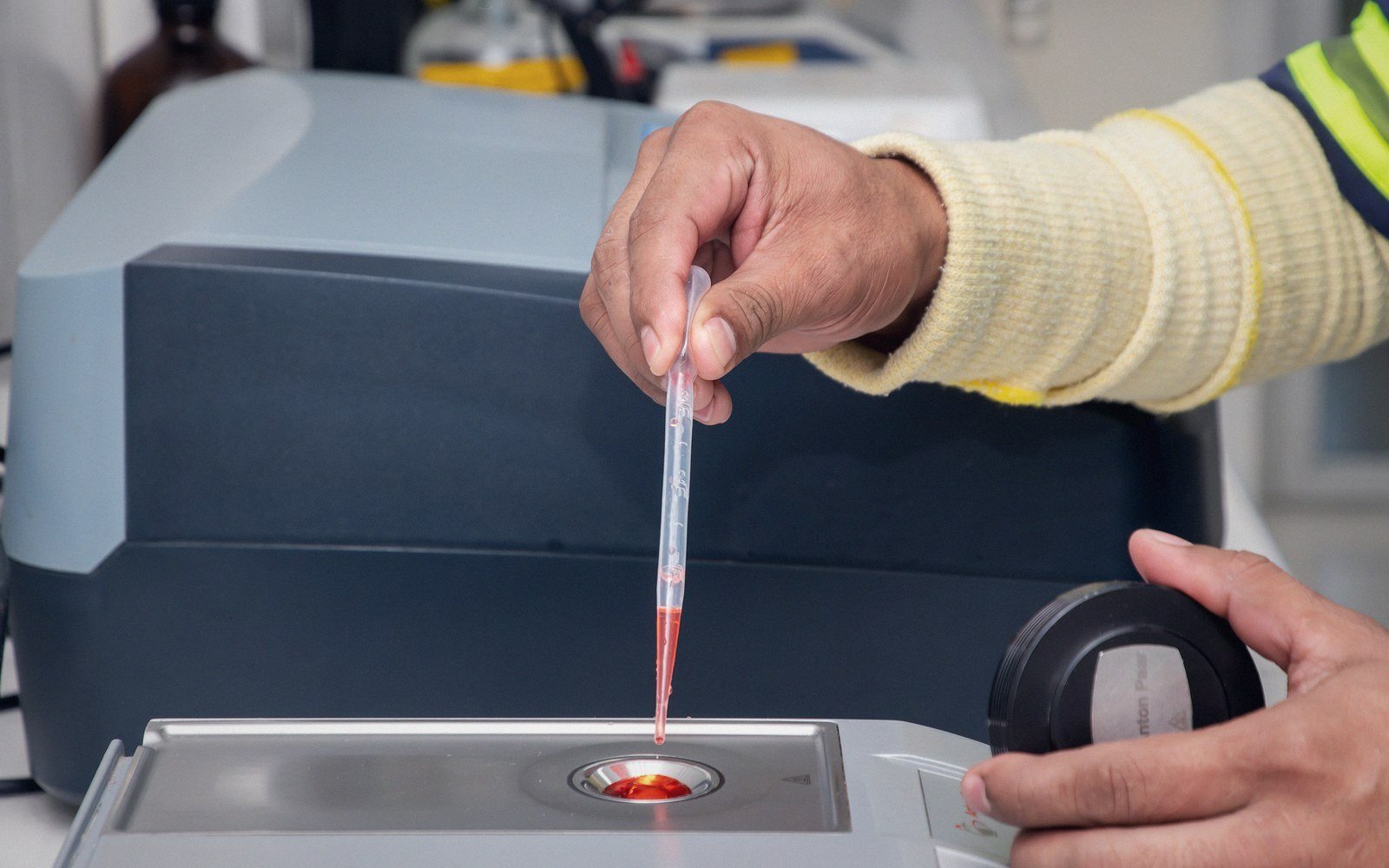 A gloved hand pipettes a red liquid sample onto a lab testing device
