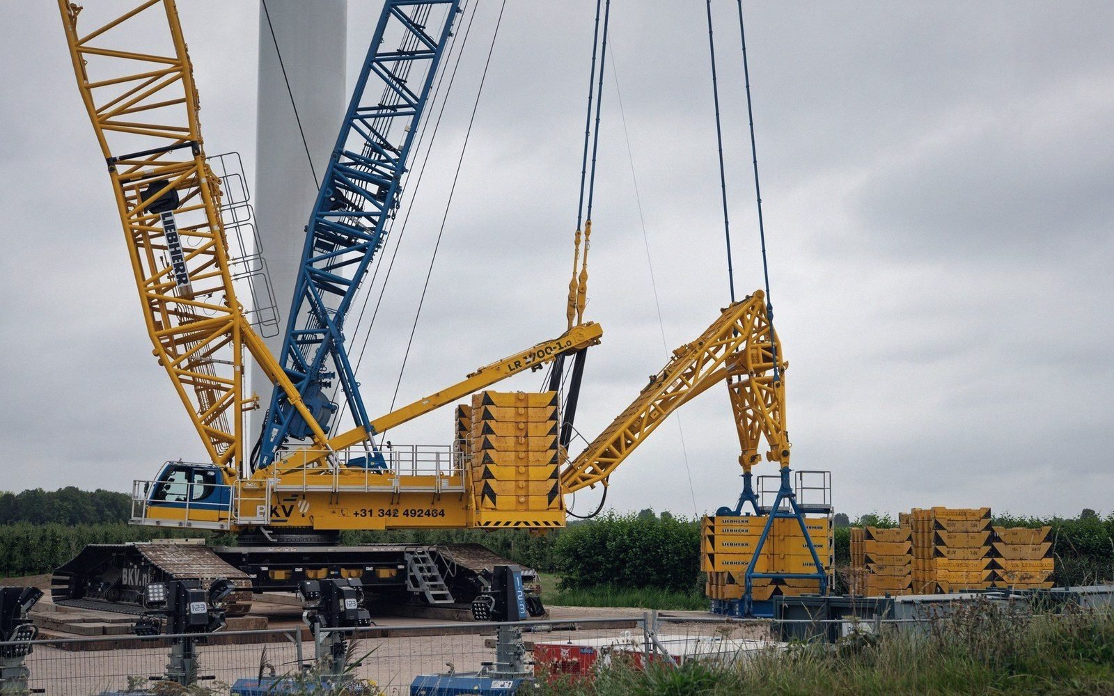 A crawler crane with counterweight blocks assembles beside a wind turbine tower