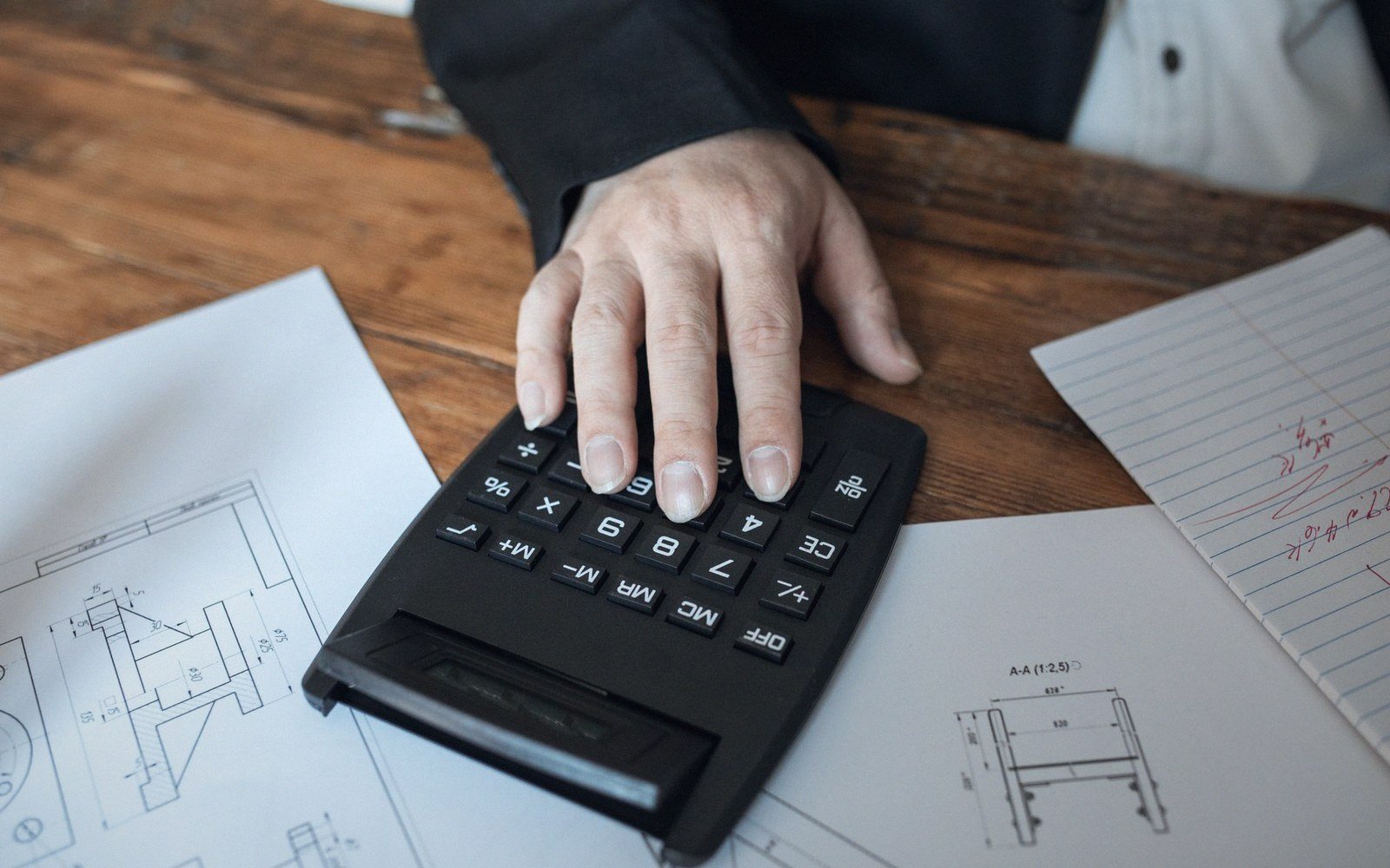 A hand pressing buttons on a calculator over engineering drawings on a desk