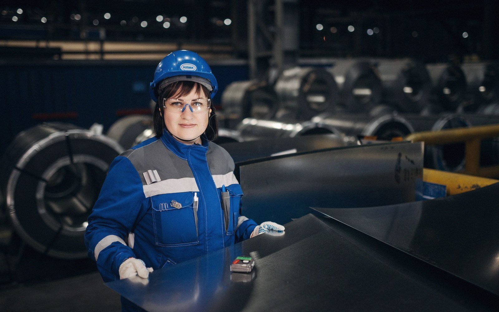 Worker in blue hard hat and safety glasses stands beside metal coils in a steel plant
