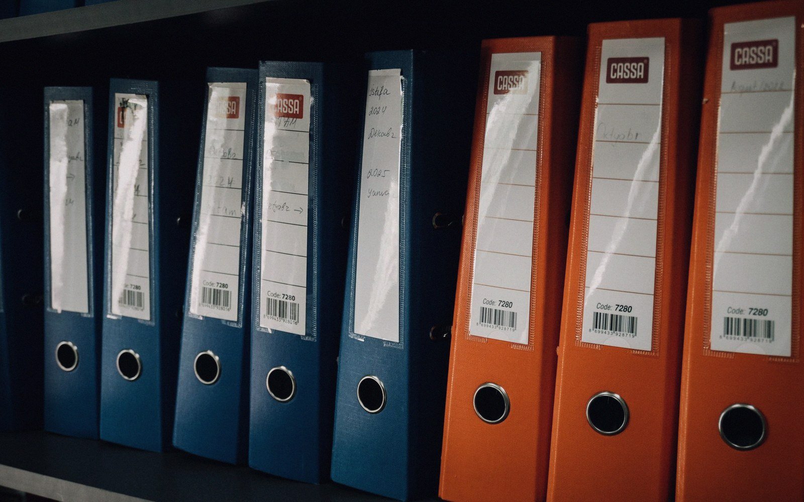 Blue and orange ring binders with handwritten labels lined up on a shelf