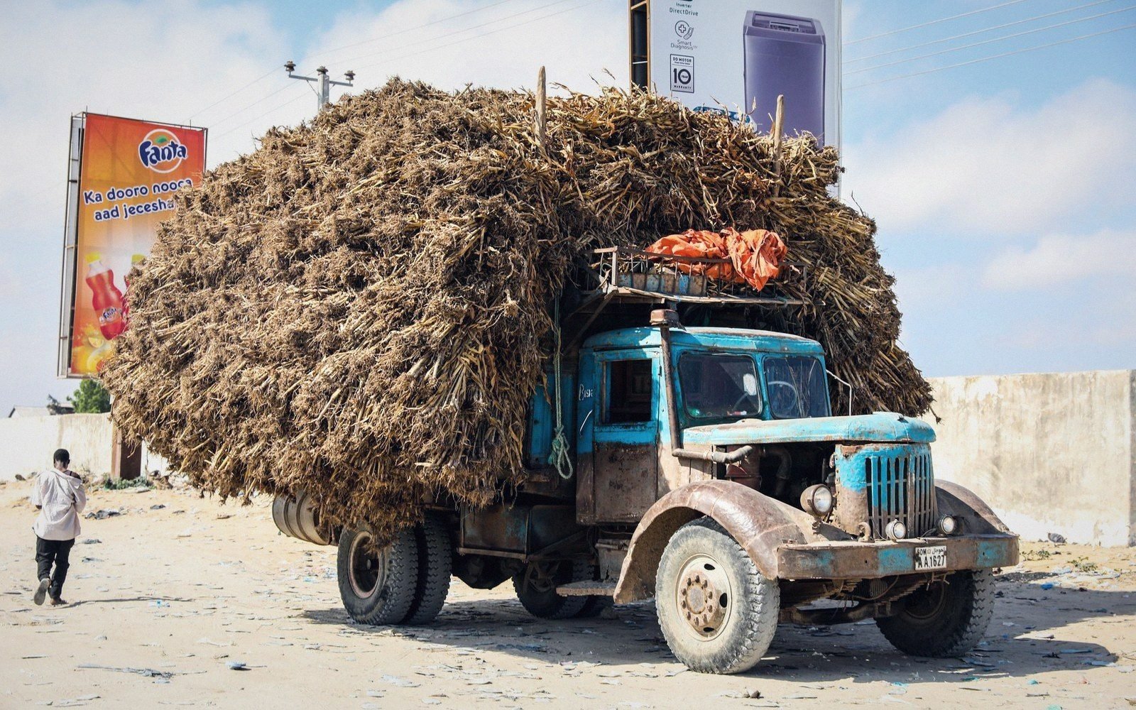 A weathered blue truck piled high with dried straw parked near roadside advertising billboards